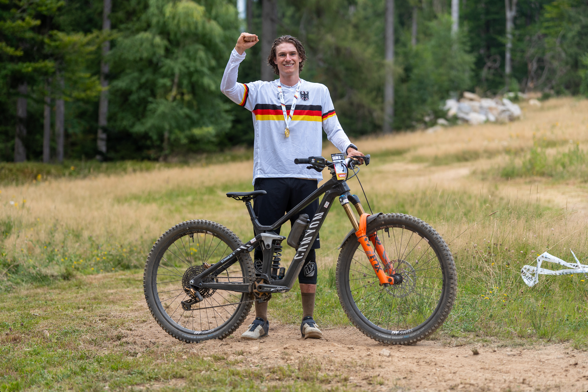 Freddy Matz wearing a white long-sleeve cycling jersey with German flag stripes (black, red, and gold) and a gold medal around his neck stands next to a black full-suspension mountain bike with orange front fork details in an outdoor grassy forest setting.
