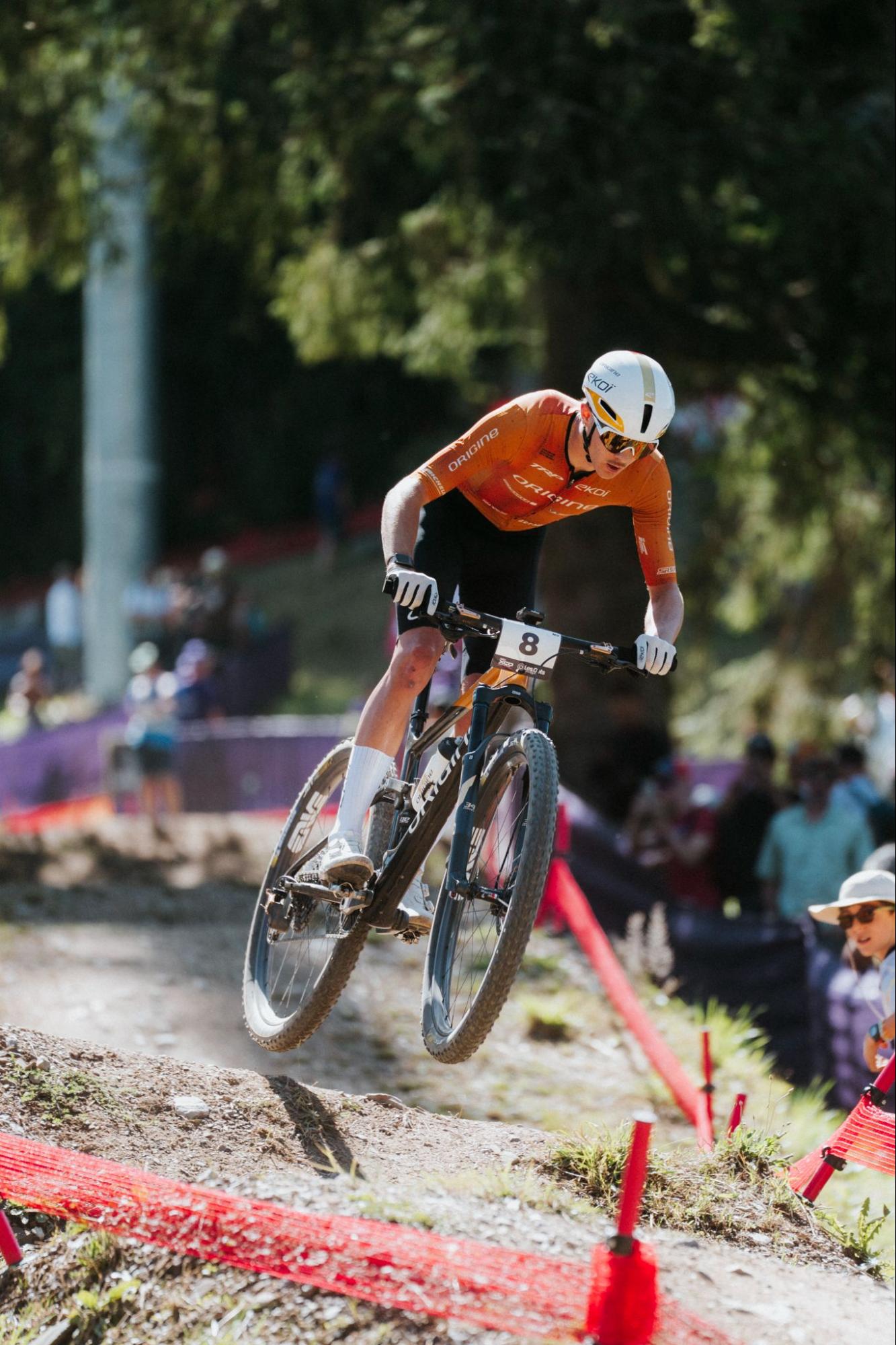 A mountain biker in an orange jersey and white helmet rides a bike over a dirt track, with trees and spectators blurred in the background.