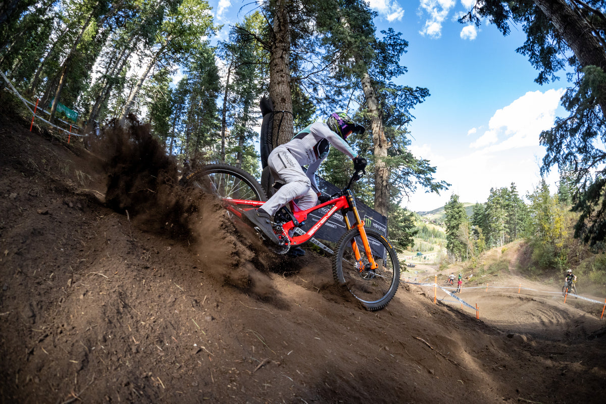 A mountain biker in white riding gear maneuvers an orange and red full-suspension bike down a dirt trail through a forest with trees, blue sky, and kicking up dust.