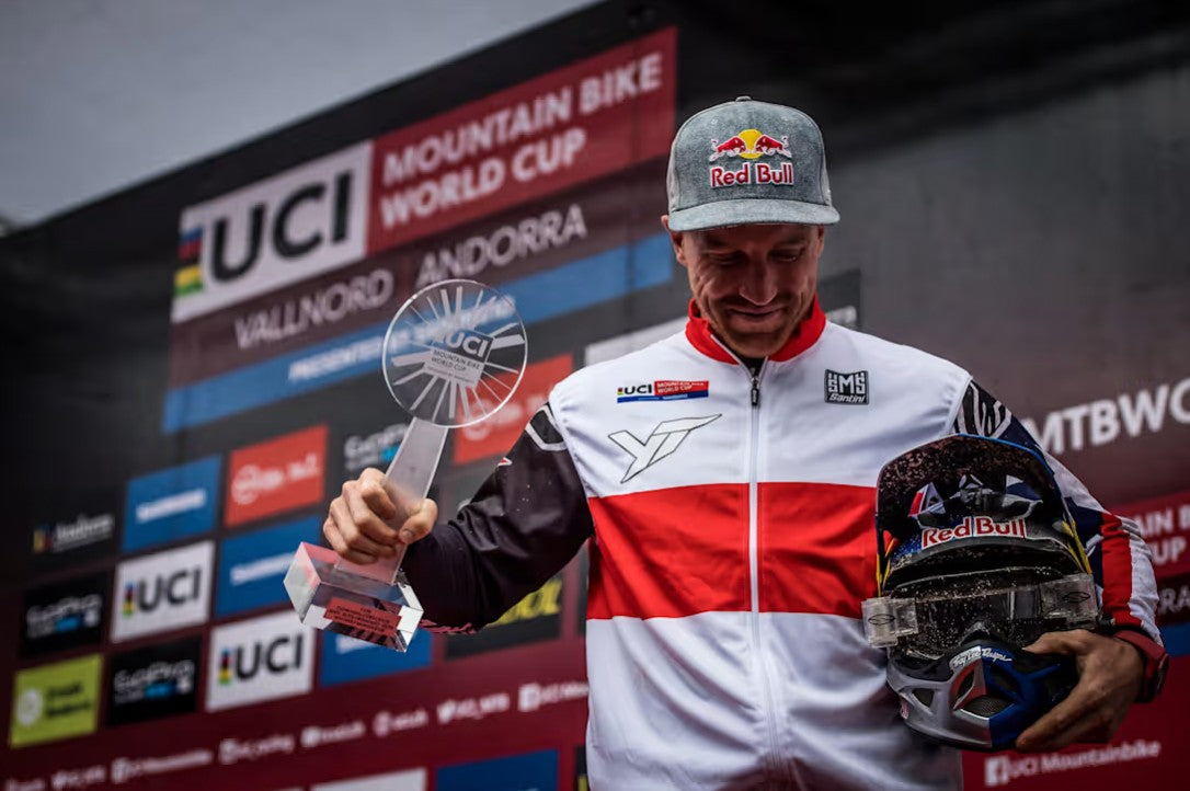 A mountain bike racer in a white, red, and black UCI World Cup jacket holds a clear trophy and a helmet on a podium in front of a UCI Mountain Bike World Cup backdrop.