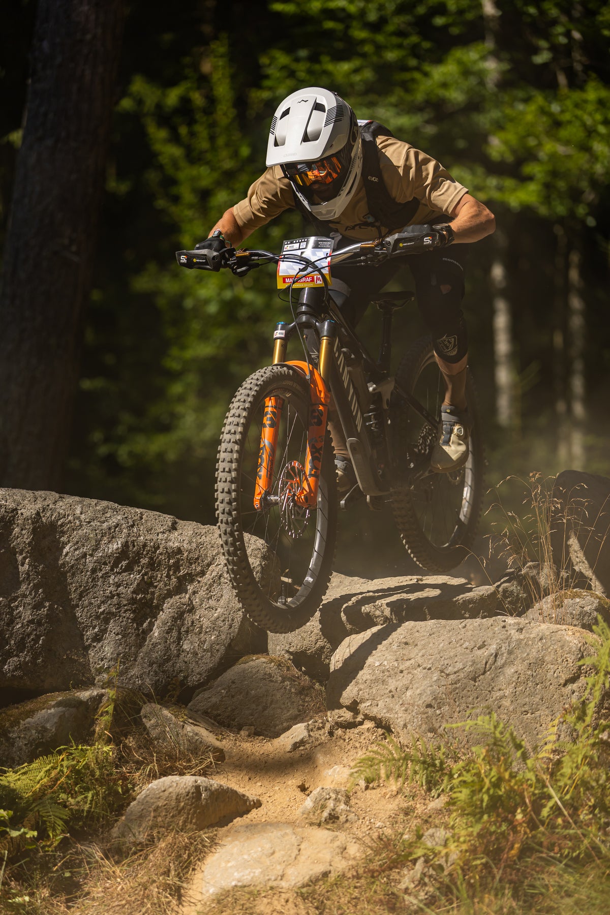 A mountain biker wearing a white helmet and tan shirt navigates a rocky forest trail on an orange and black full-suspension bike.