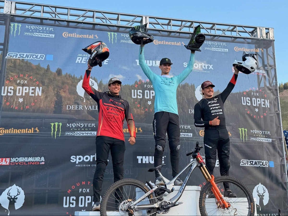 Three mountain bikers stand on a podium holding trophies and helmets in front of a large sponsor backdrop. A mountain bike is propped in the foreground, and the scene is outdoors under daylight.