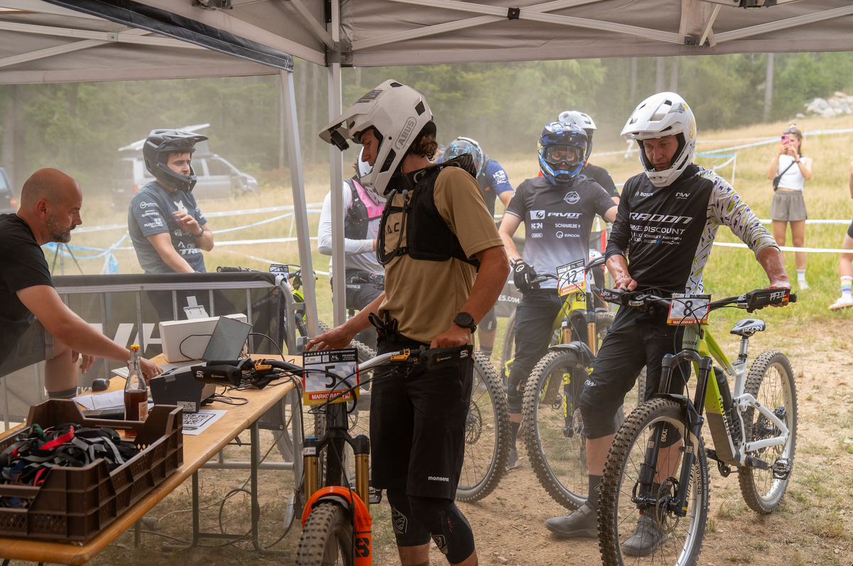 Mountain bikers wearing helmets and protective gear gather under a white canopy tent at a bike event, with one rider in a black jersey standing next to a bright green bike.