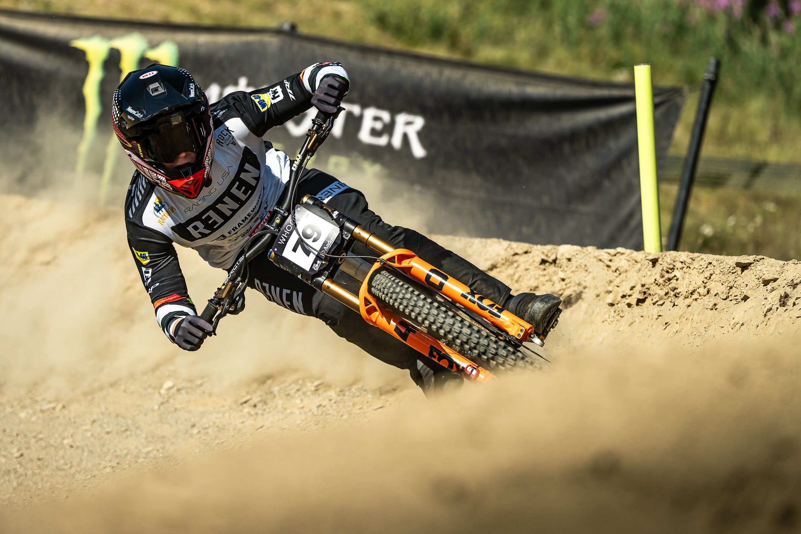 A mountain biker in a black and white racing suit leans sharply into a dirt track turn, kicking up dust. The bike is orange and black, and a black Monster Energy banner is visible in the background.