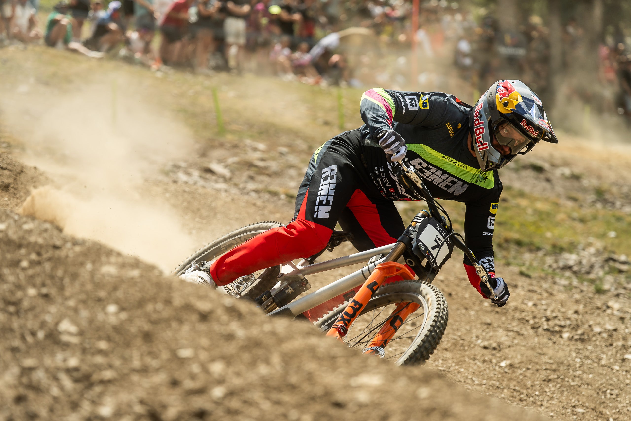 A mountain biker in a black, red, and green racing suit leans sharply into a turn on a dirt track, kicking up dust. The rider wears a Red Bull helmet and the brand name 'Red Bull' is visible on the helmet and suit.