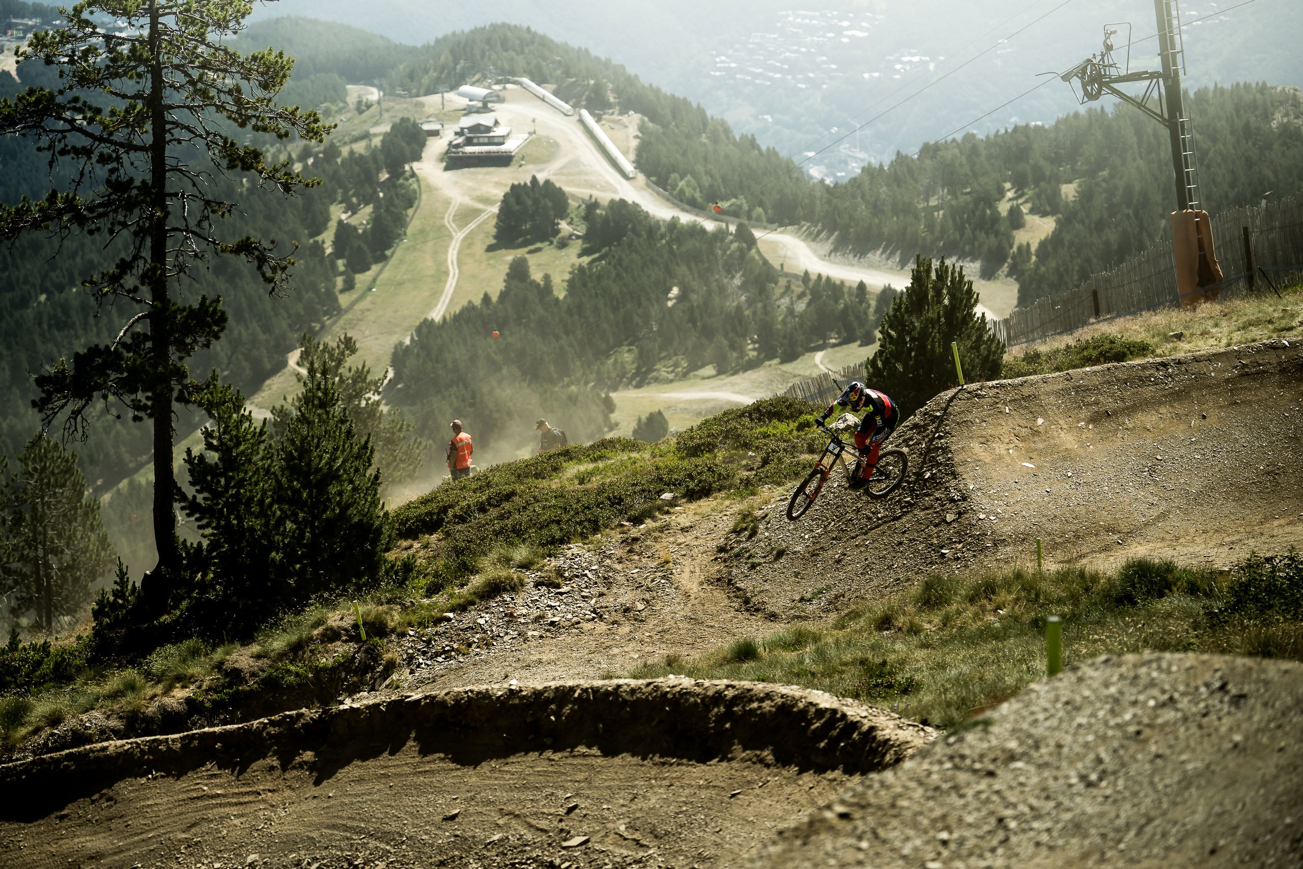 A mountain biker navigates a dirt jump track on a steep mountainside, with a ski resort and forested hills visible in the background.