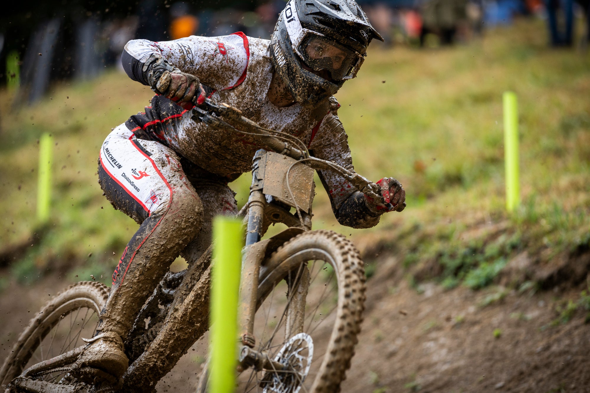 A muddy mountain biker wearing a full-face helmet and goggles leans into a turn on a dirt bike over a muddy track, with bright yellow markers and a blurred grassy background.