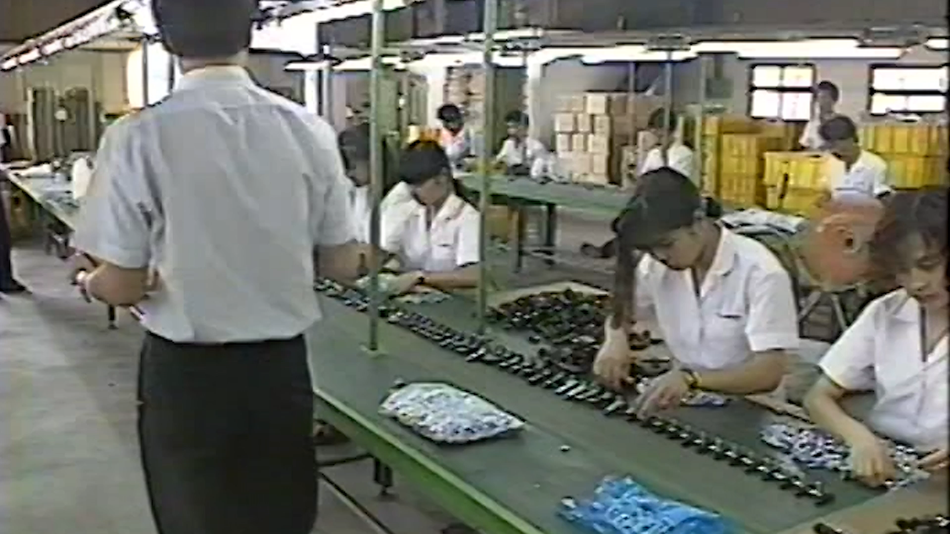 A factory floor scene with several workers in white shirts assembling or inspecting long rows of small parts on a green conveyor table. A person in the foreground stands with their back to the camera, and boxes are stacked in the background.