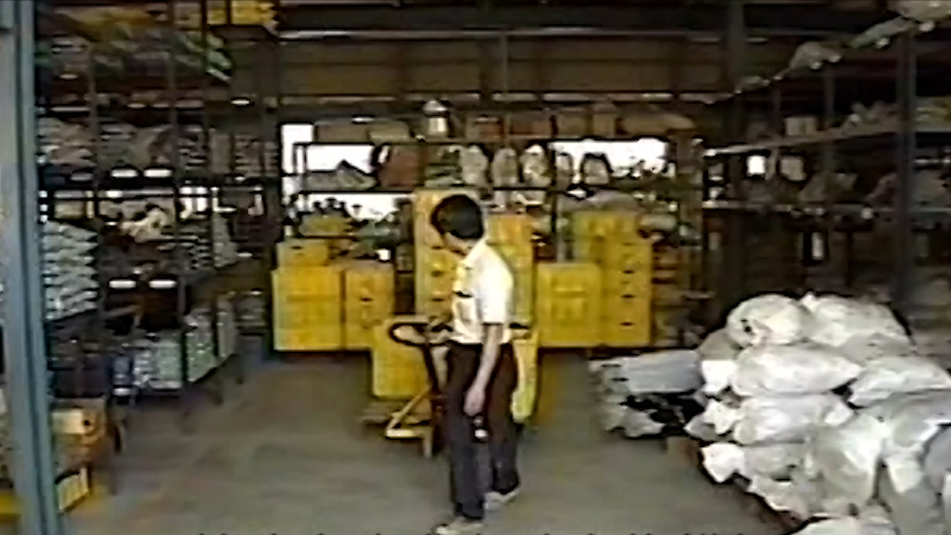 A person in a warehouse standing next to a yellow pallet jack, surrounded by shelves filled with goods and a large pile of white bags on the right.