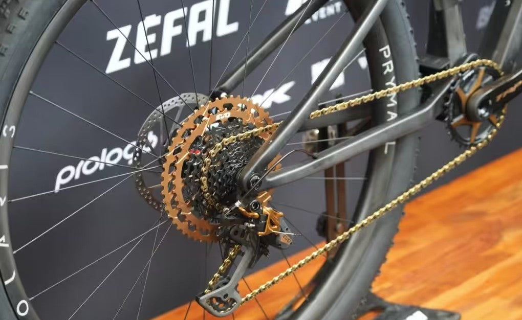 Close-up of a bicycle rear wheel and drivetrain, showing a black frame, black tire, gold chainring, gold chain, and a dark branded backdrop with ZEFAL and prisma logos.