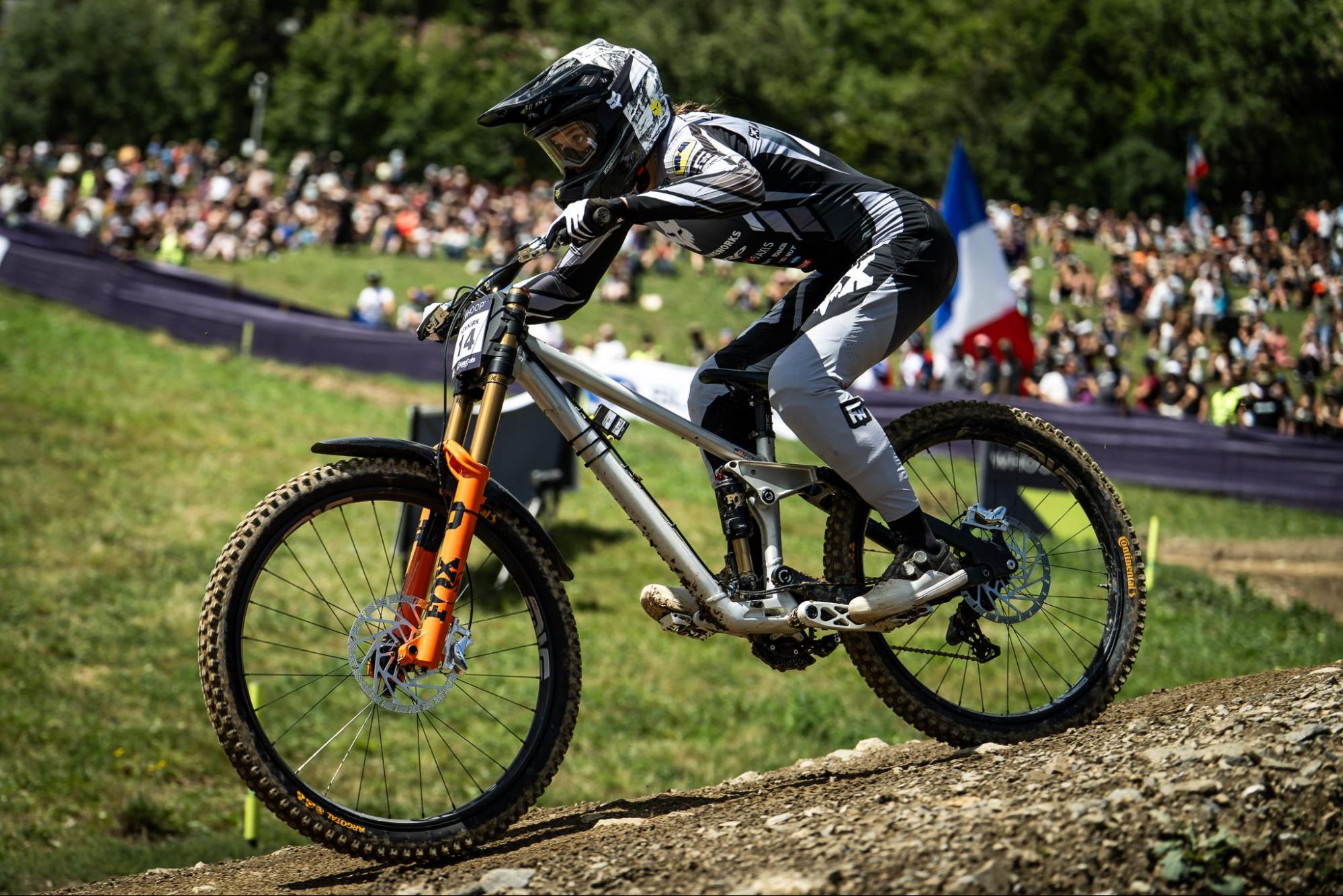 A mountain biker in a black and white racing suit leans sharply over a dirt jump, riding a silver full-suspension bike with an orange front fork on an outdoor track with a crowd in the background.