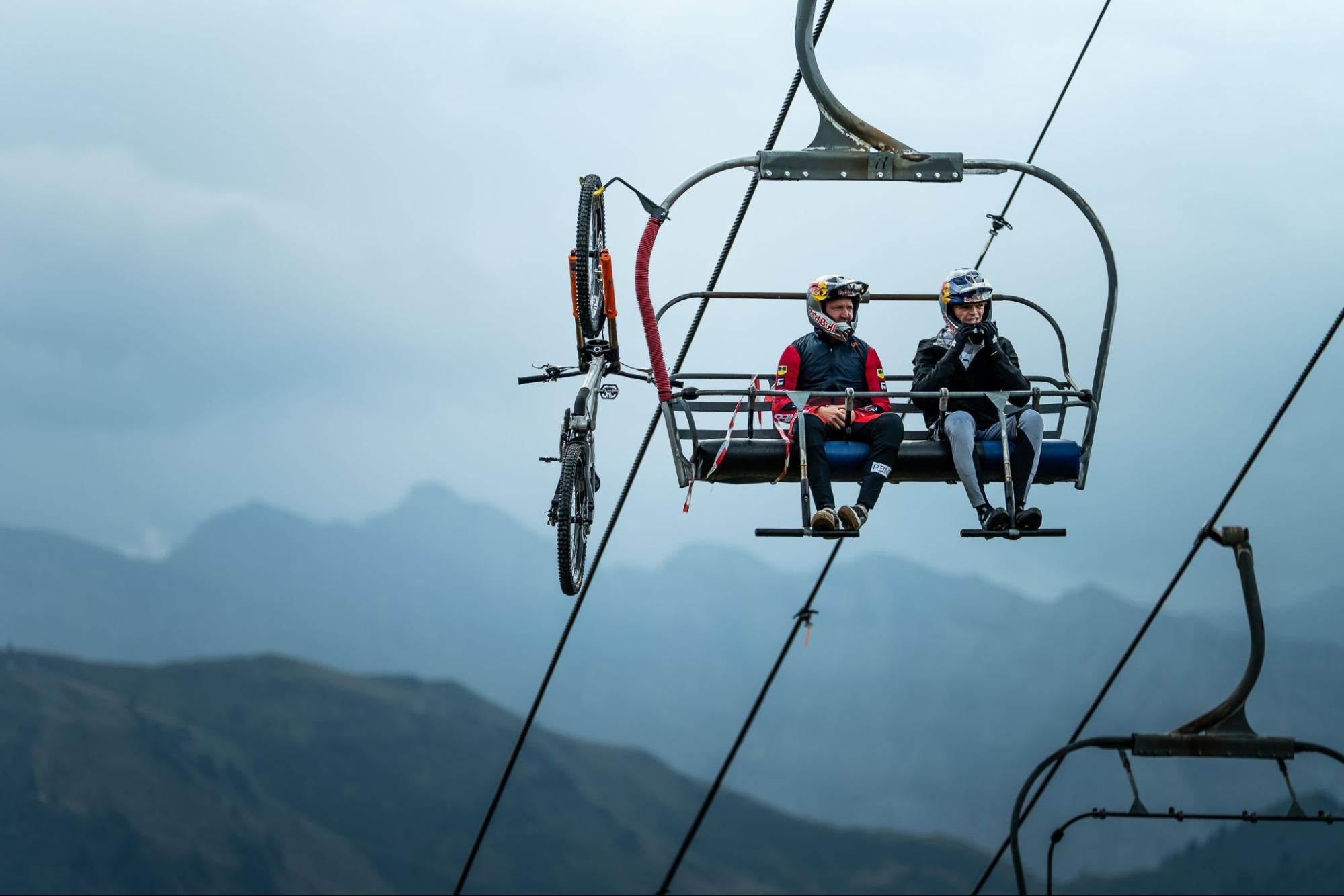Two mountain bikers wearing helmets sit on a ski resort chairlift with a mountain range in the background under an overcast sky.