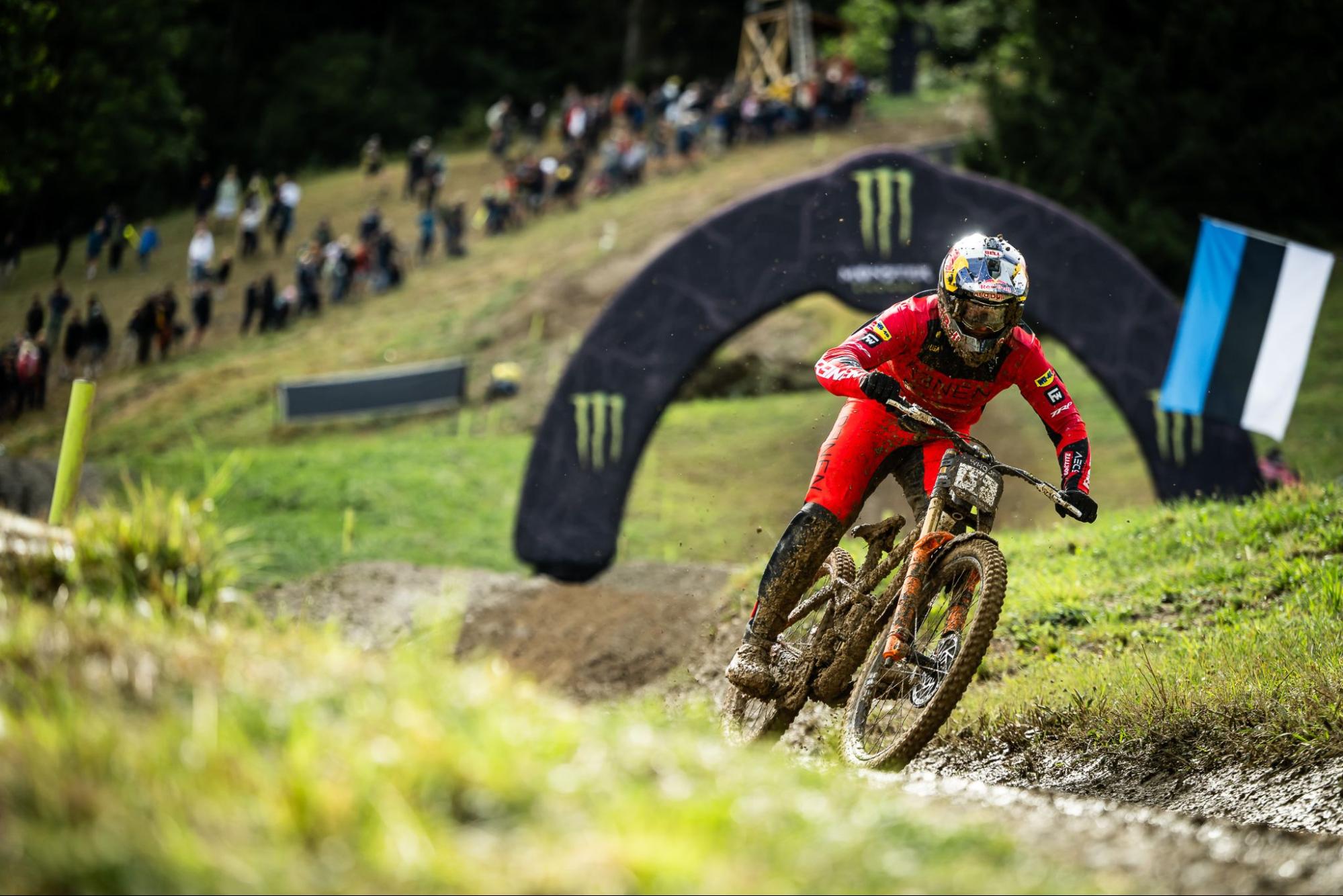A mountain biker in a red racing suit and full-face helmet navigates a dirt track turn, with a blurred grassy foreground and a large black Monster Energy archway in the background.