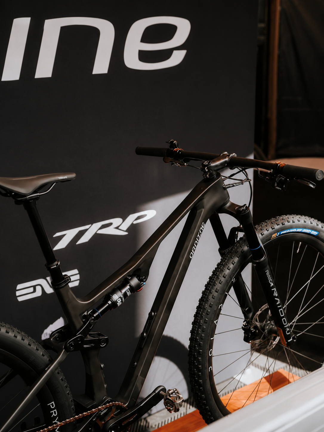 A black full-suspension mountain bike with a matte black frame and knobby tires, displayed in front of a black backdrop featuring the white TRP logo.