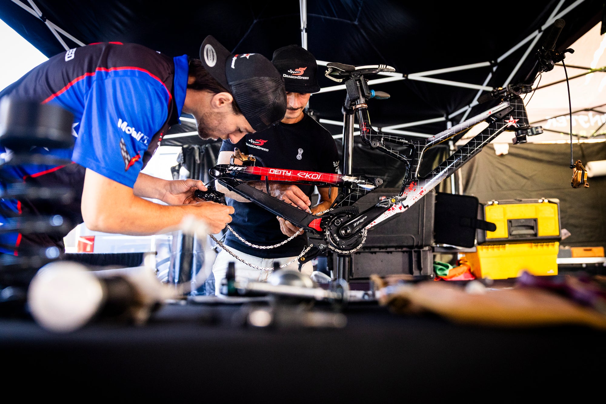 A mountain bike is mounted on a work stand inside a tent while two mechanics in branded blue and black jerseys work on the bicycle frame.