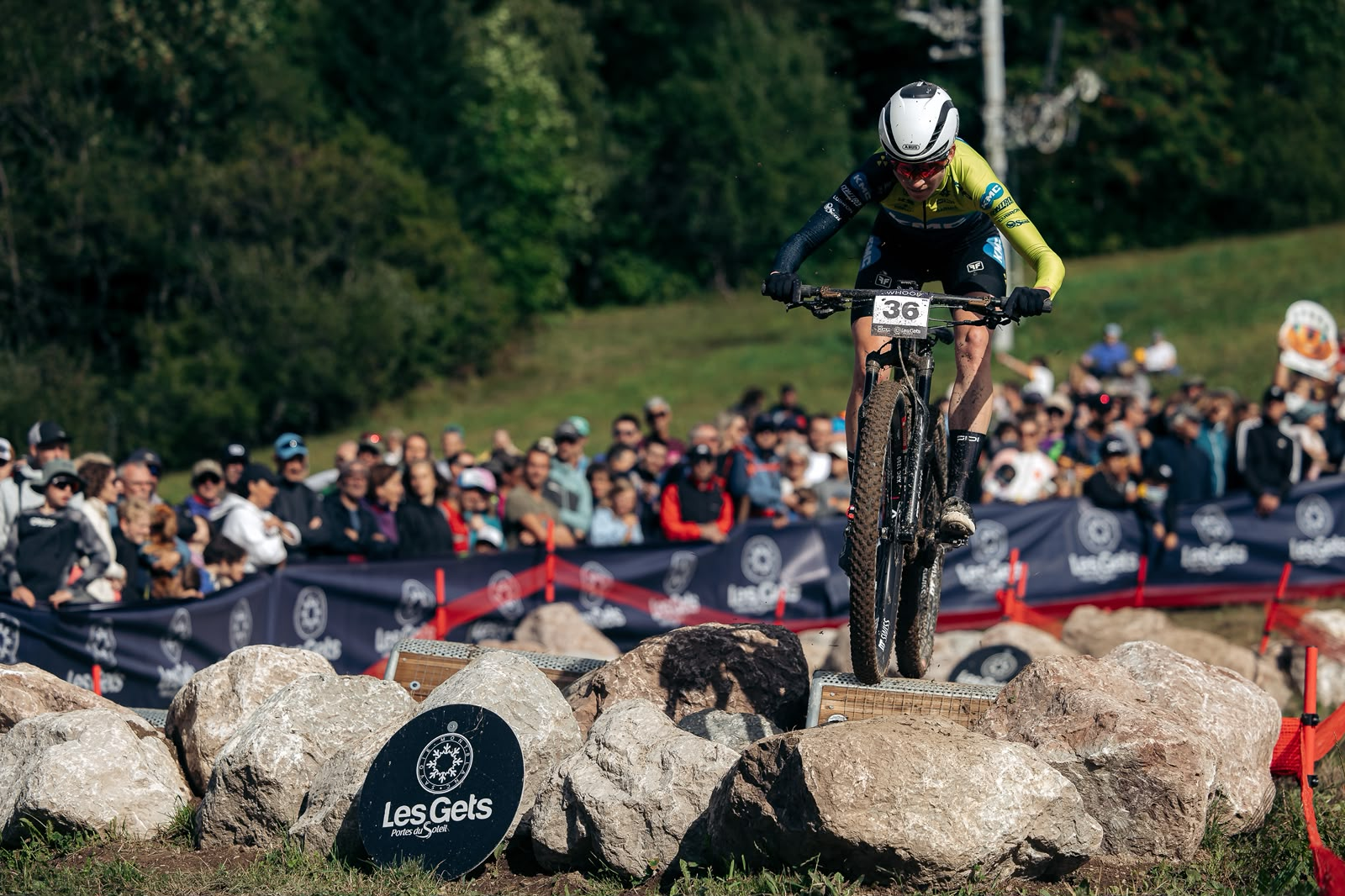 A mountain biker wearing a white helmet and a green, black, and blue jersey rides a bike over a rocky obstacle at an outdoor race, with a crowd of spectators in the background.