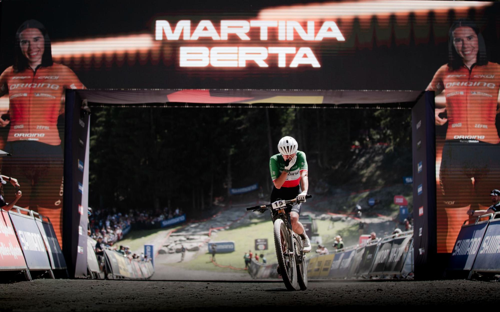 A mountain biker wearing a green, white, and red jersey rides through a large archway with 'MARTINA BERTA' displayed prominently above, set against a dark forest backdrop.