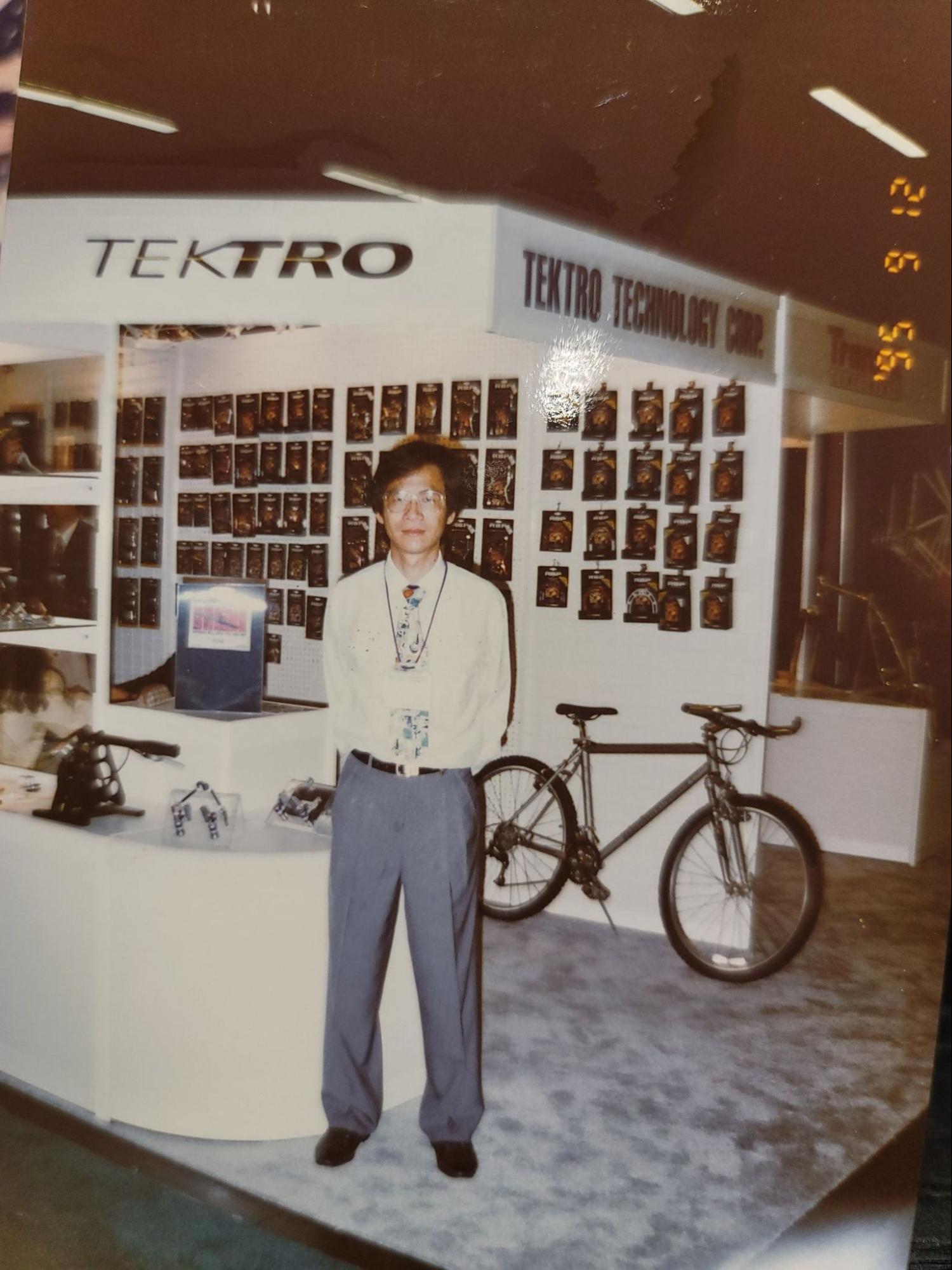 A vintage photograph showing a man standing in front of a TEKTRONICS booth at an exhibition, with a bicycle parked next to him. The photo has a warm, aged color cast and a digital timestamp in the top right corner.