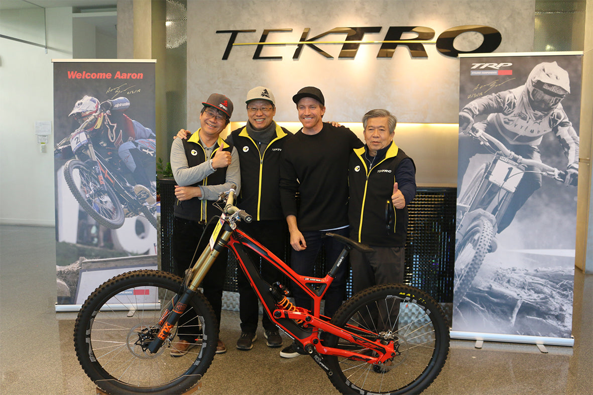 Four men posing with a red full-suspension mountain bike in front of a Tektro wall display, with two men wearing matching black and yellow Tektro jackets.