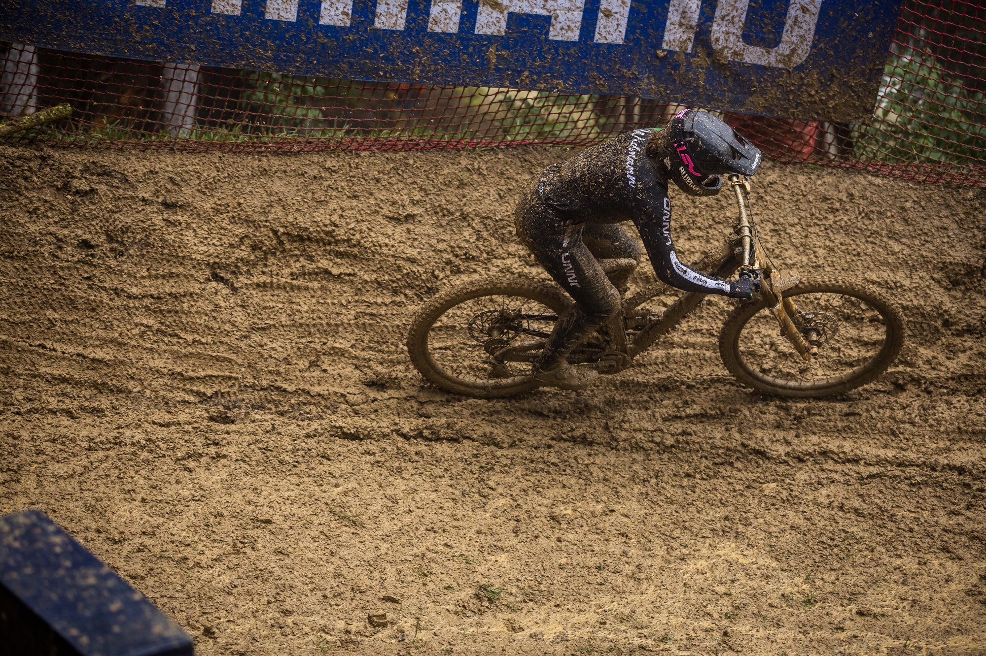 A mountain biker in a full-face helmet and muddy suit riding a dirt bike on a muddy track, with a blue barrier and chainlink fence in the background.