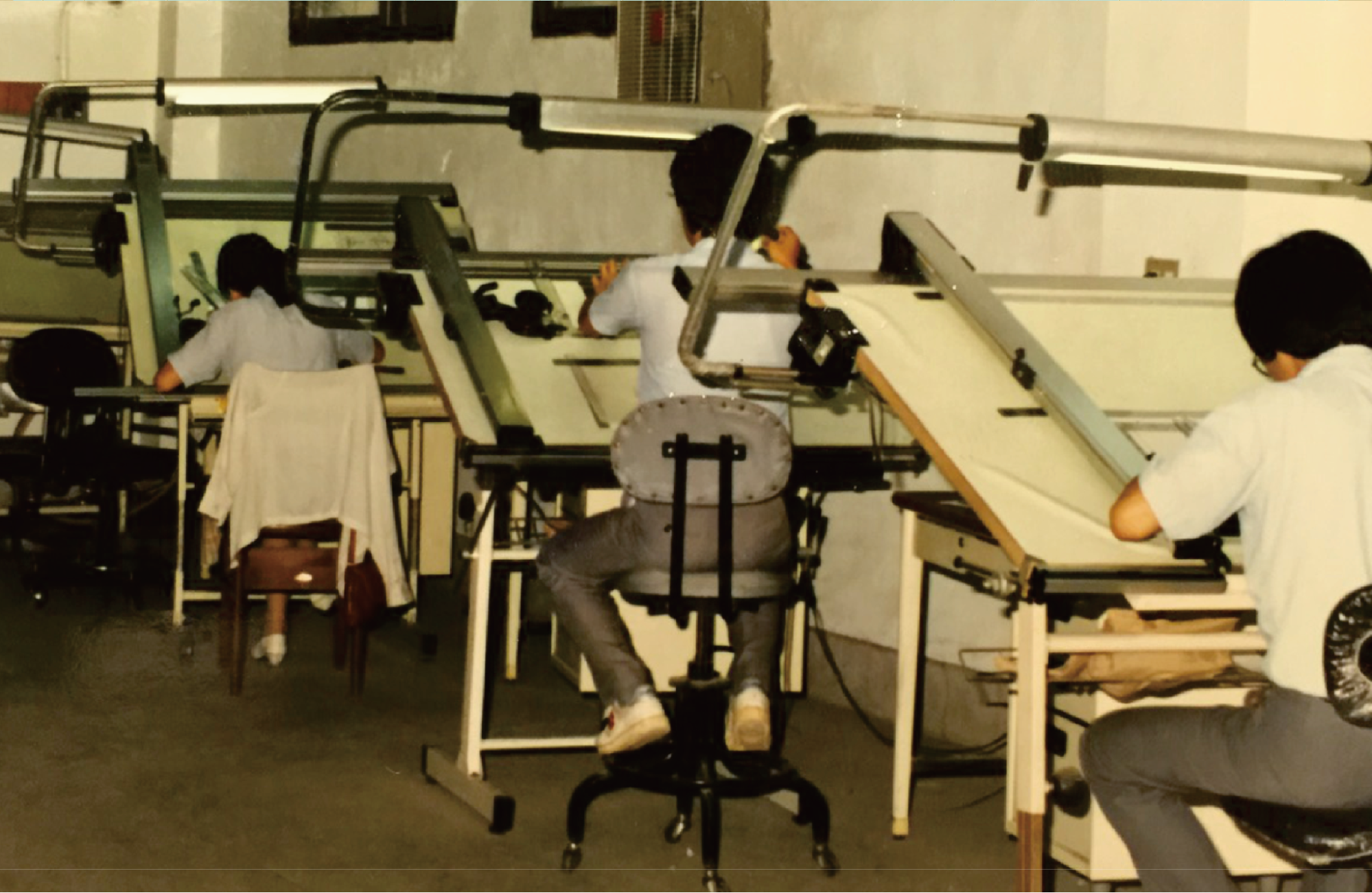 A vintage photograph showing several people seated at large drafting or printing machines in an indoor workshop or studio. The scene includes office chairs, tables, and long metal drafting arms, with one person centered in the foreground facing away from the camera.