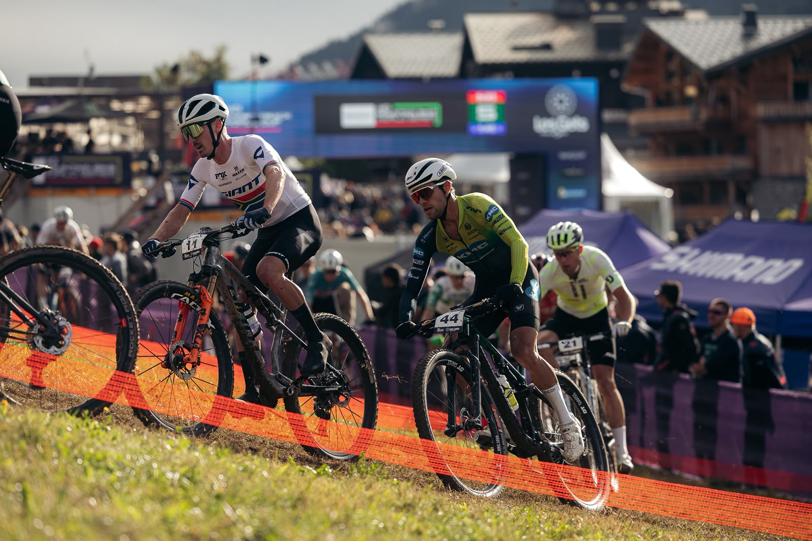 Mountain bike racers compete on a grassy downhill course during an outdoor cycling event, with spectators and race tents in the background.