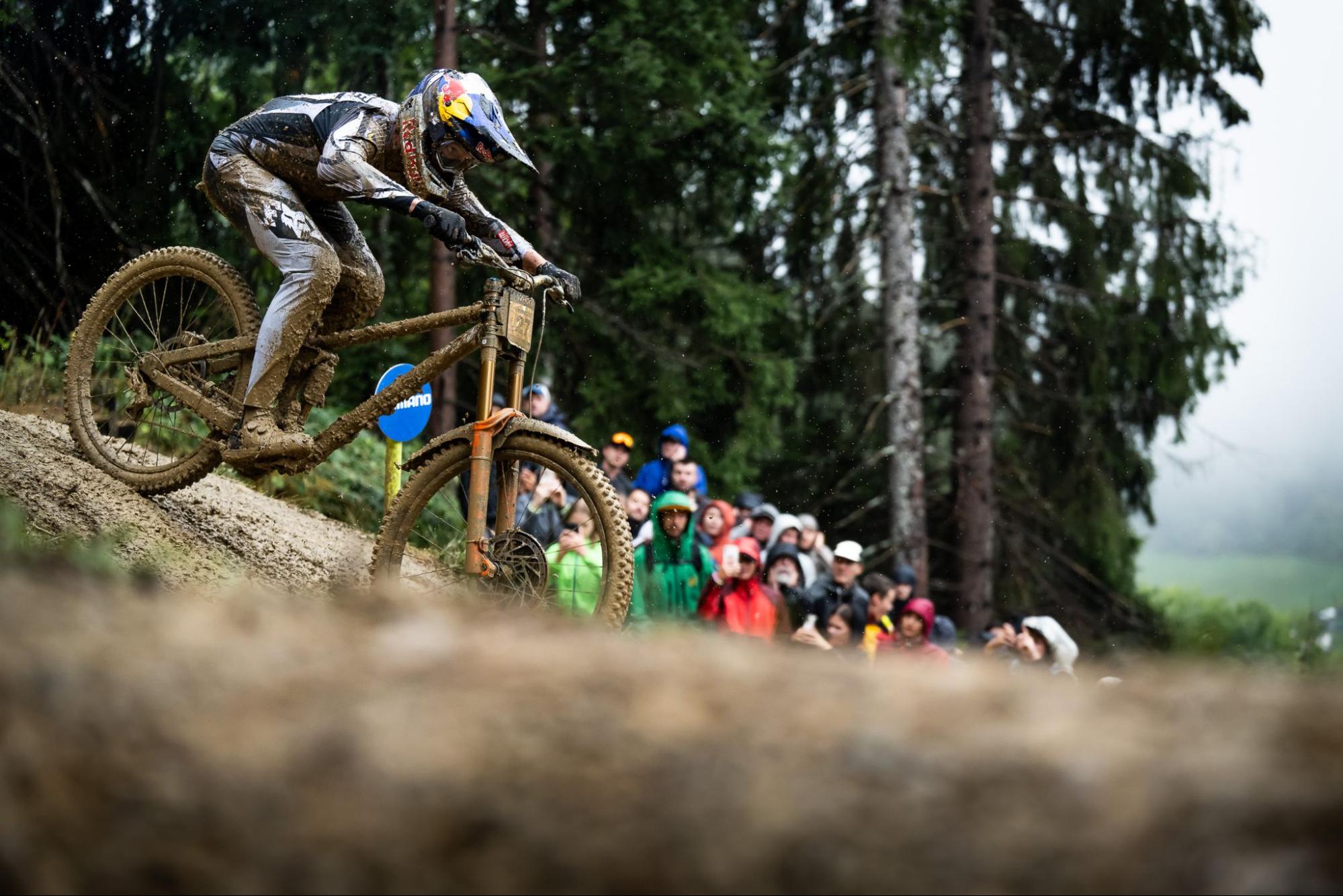 A full-face mountain biker in muddy gear leans sharply on a dirt bike during an outdoor race, with a blurred crowd of spectators and tall evergreen trees in the background.