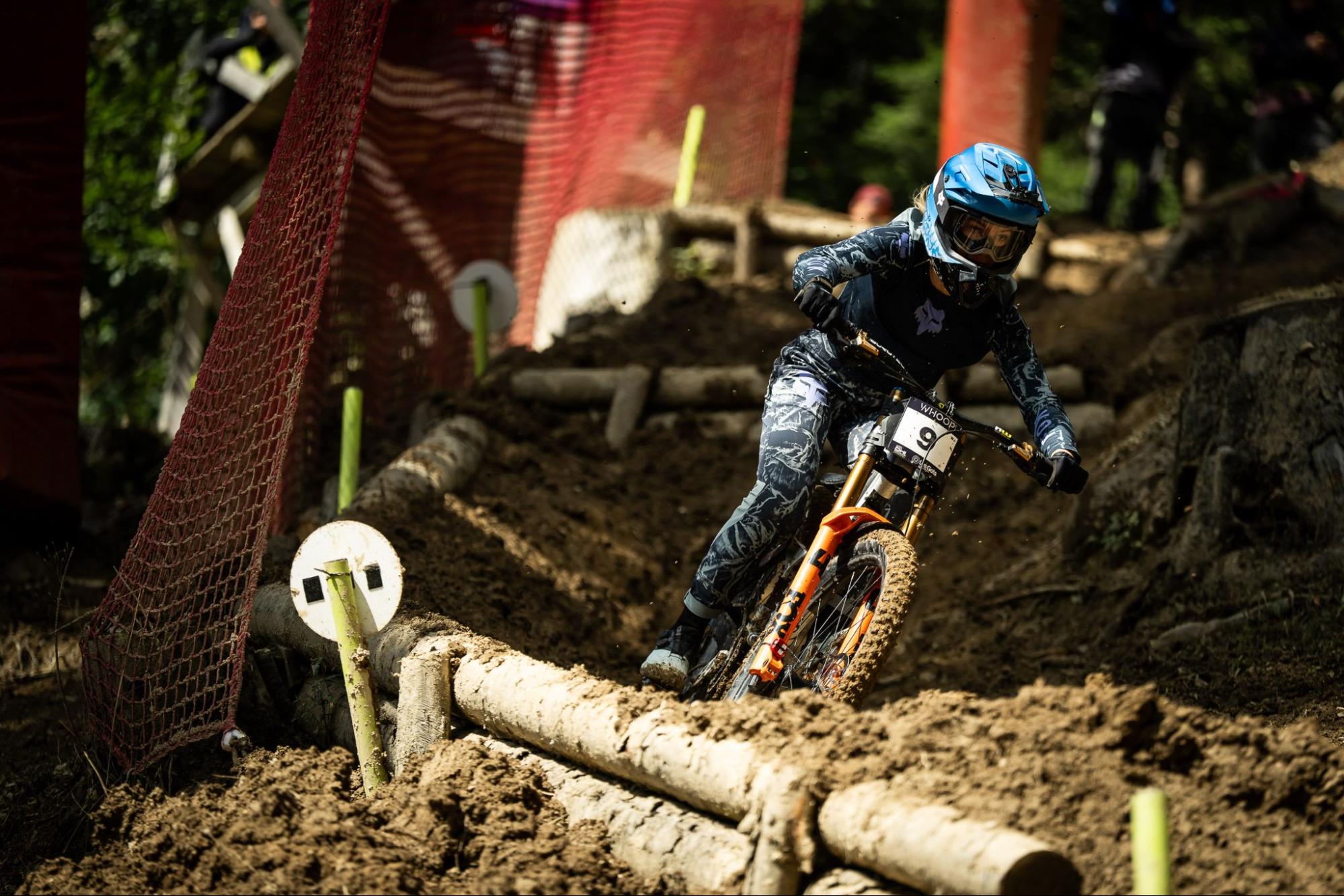 A mountain biker wearing a blue helmet and dark patterned riding suit navigates a muddy dirt track with logs and safety netting visible in the background.