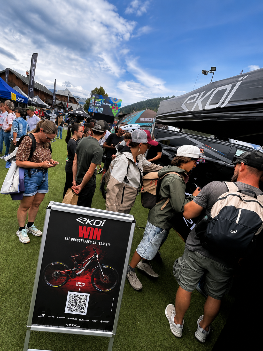 Outdoor event scene with multiple people gathered around a black canopy tent and a freestanding sign that reads 'ekoi WIN'. The setting is on grass under a blue sky, with tents and other booths visible in the background.