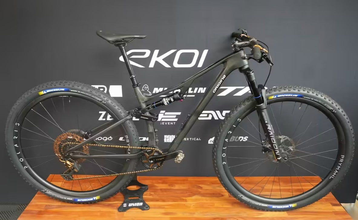 A black full-suspension mountain bike displayed on a wooden stand against a dark gray branded backdrop. The bike frame shows the Micron logo, with visible components including a suspension fork, knobby tires, disc brakes, and a rear suspension spring.