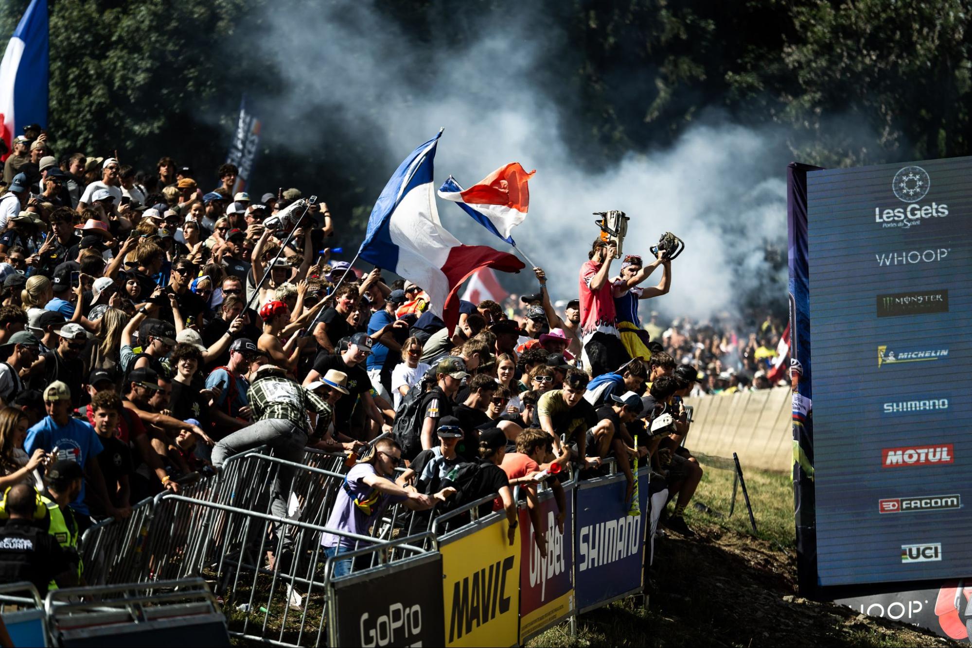 A crowded outdoor event scene with a large group of spectators behind metal barriers, smoke in the background, and several people holding French flags. A large sponsor board on the right displays brands including LesGels, Whoop, Michelin, Shimano, and Motul.