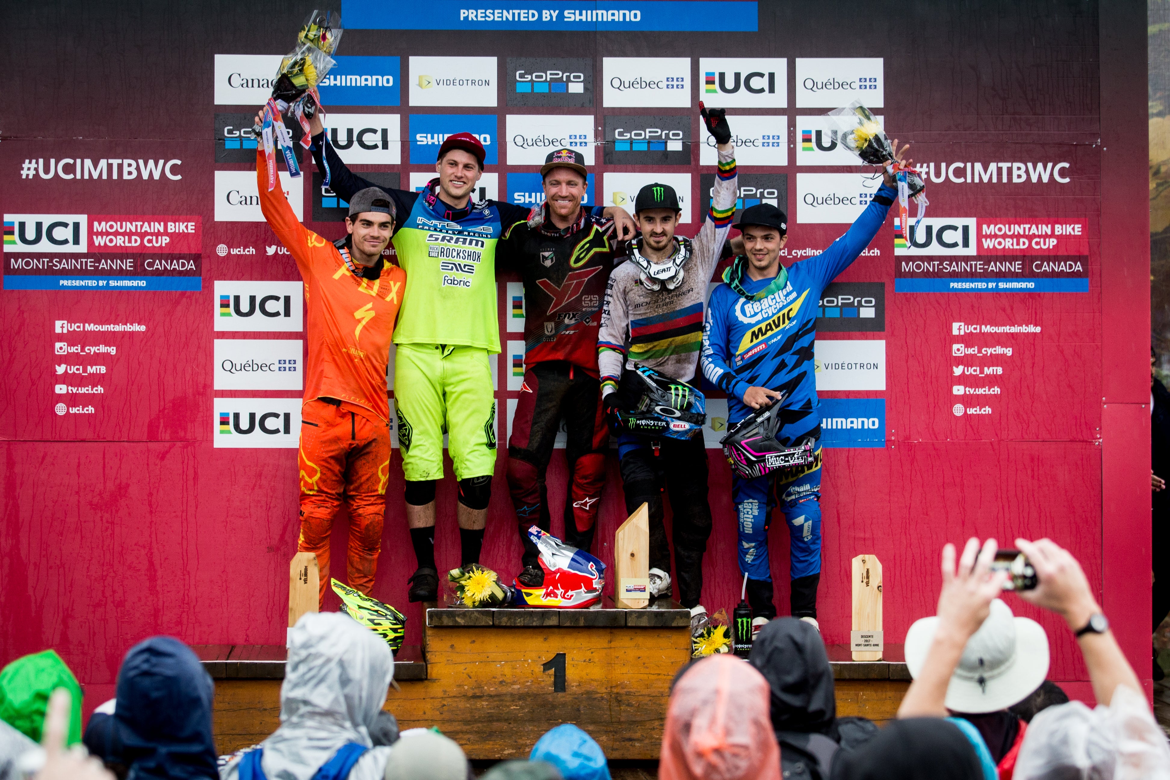 Four mountain bike racers stand on a podium holding champagne flutes in front of a UCI Mountain Bike World Cup backdrop. The racers wear colorful racing suits: orange, neon green, black, and blue with rainbow stripes.